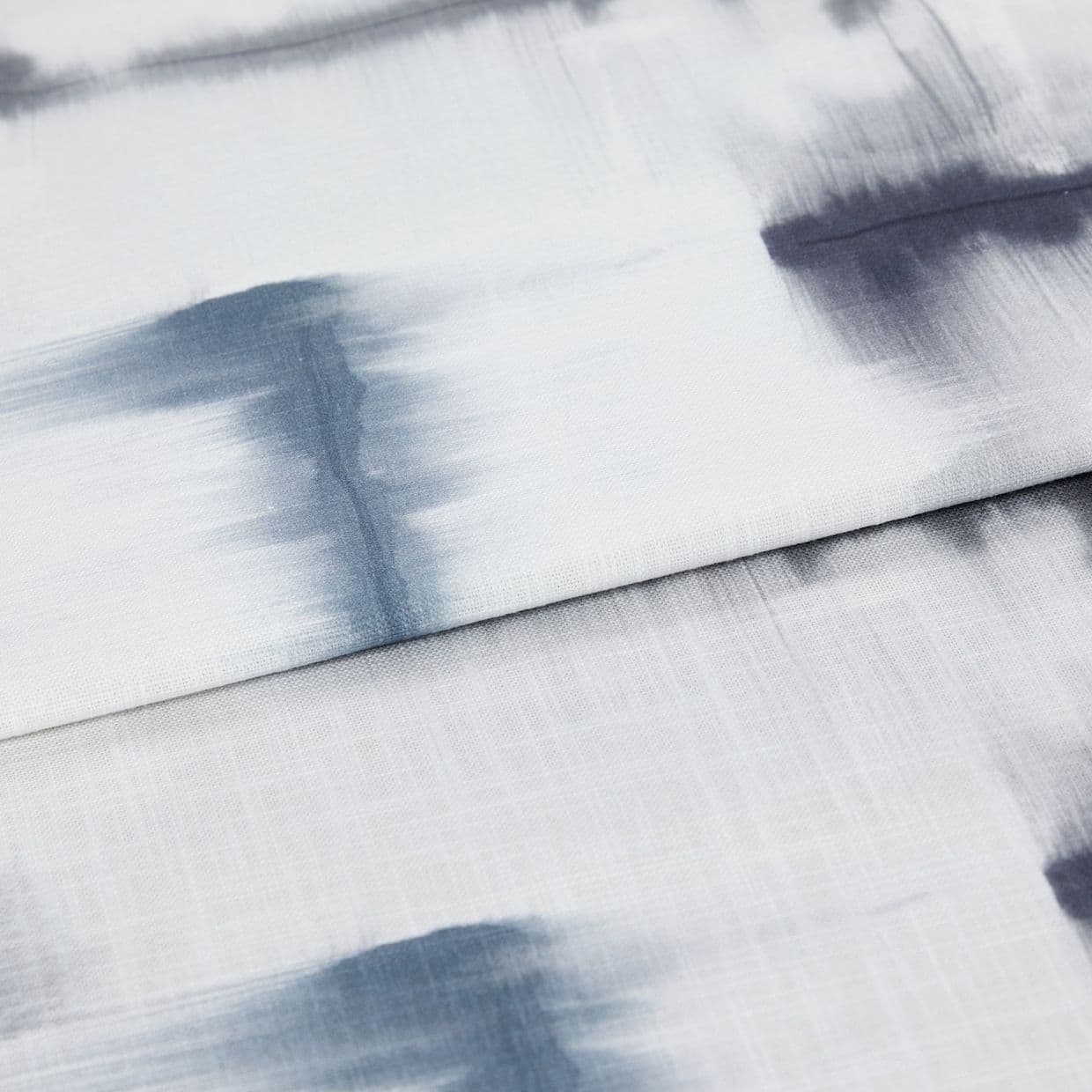White linen curtain with horizontal folds and diffuse blue-gray brushstroke pattern; folded panel overlapping below, draped to show textured weave, photographed close-up under soft, diffuse lighting.