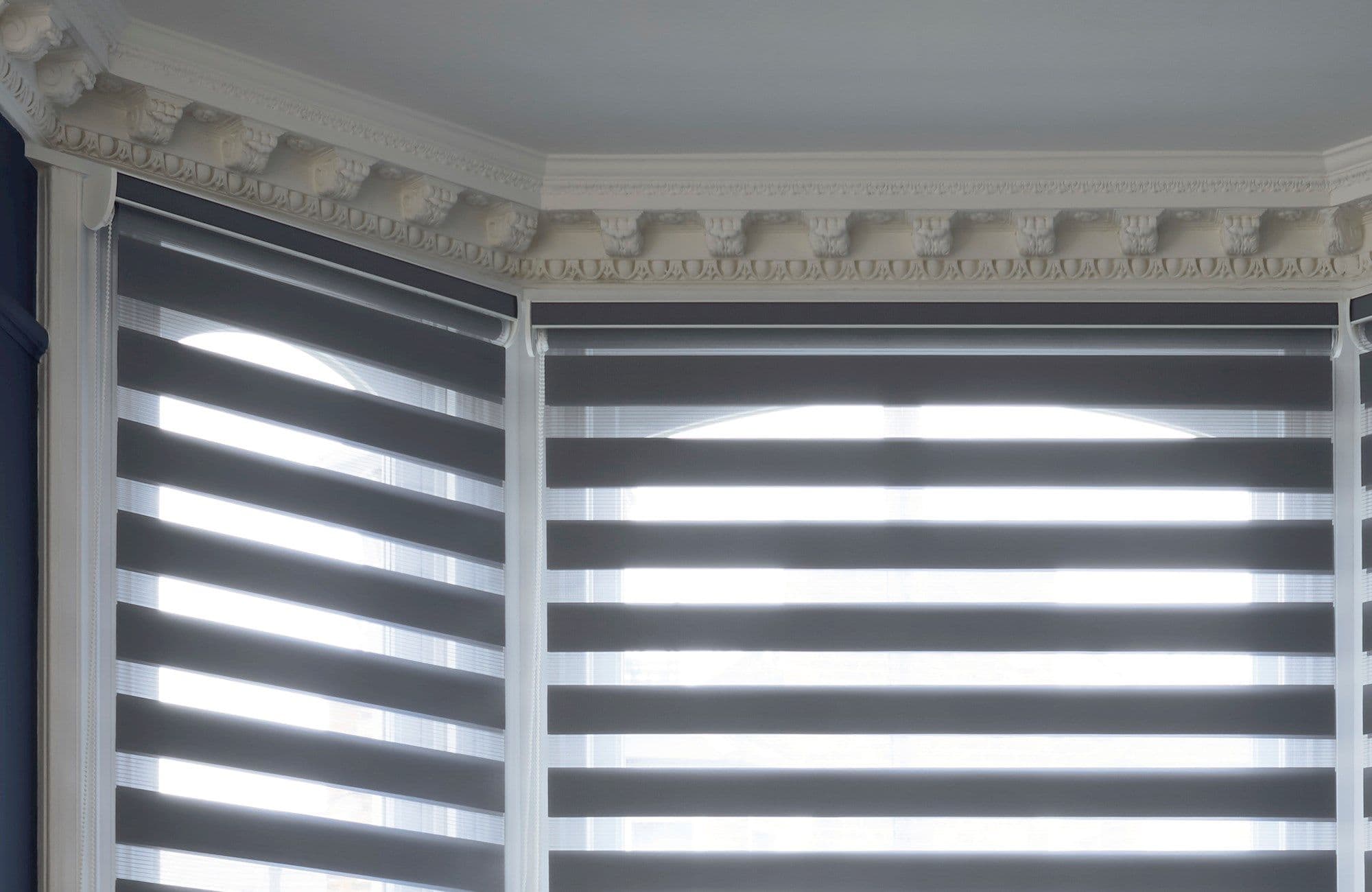Layered zebra roller blinds cover bay windows, alternating dark opaque and sheer horizontal bands, partially lowered and filtering strong daylight; ornate white cornice molding tops the bright interior.