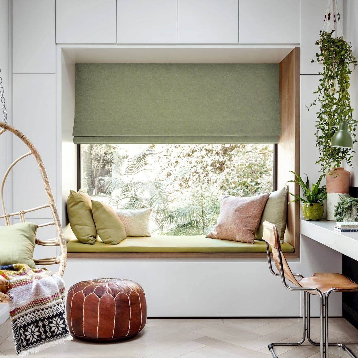 Green fabric Roman blind, lowered to cover the upper window, softly filtering daylight; framed bay window with cushioned bench, assorted pillows, hanging plants, desk and chair, bright leafy garden view.