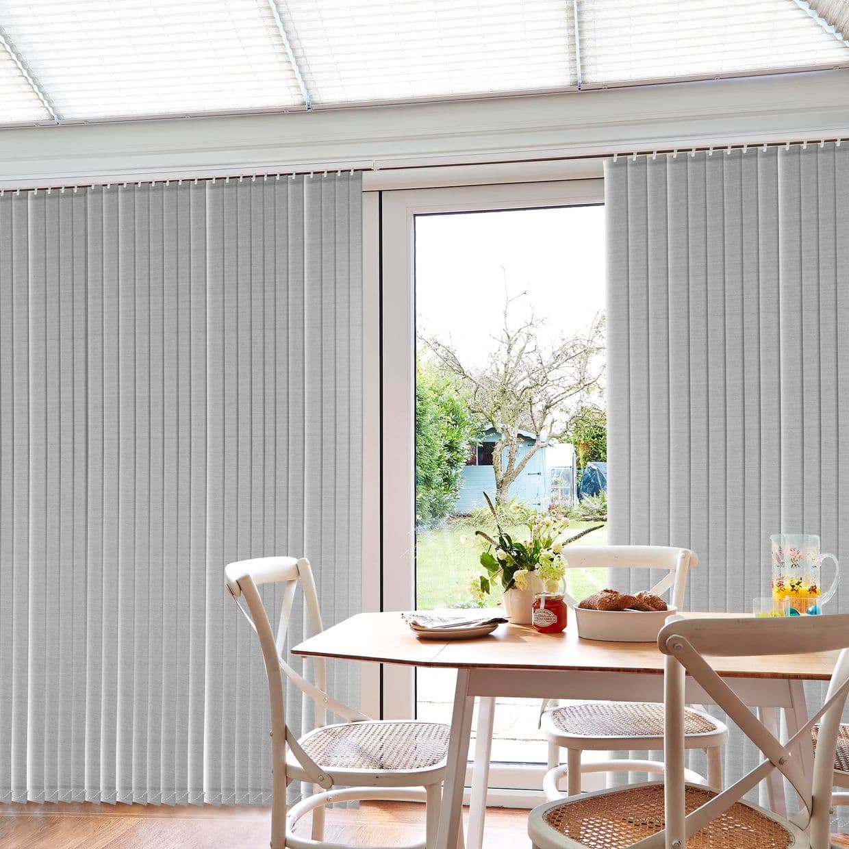 Grey ceiling-mounted vertical fabric blinds with narrow slats, drawn partly aside to reveal a central glass door, in a bright dining area with wooden table, chairs, vase and garden view.
