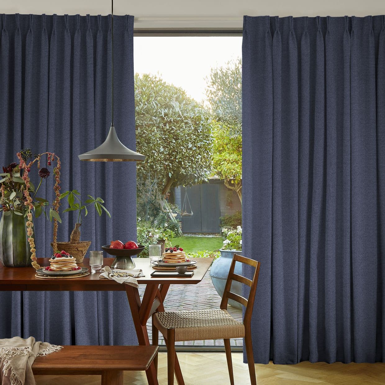Floor-length navy-blue pleated curtains, drawn open at center to reveal a window; framing a sunlit garden beyond while a wooden dining table with pancakes and a pendant lamp fills the foreground.