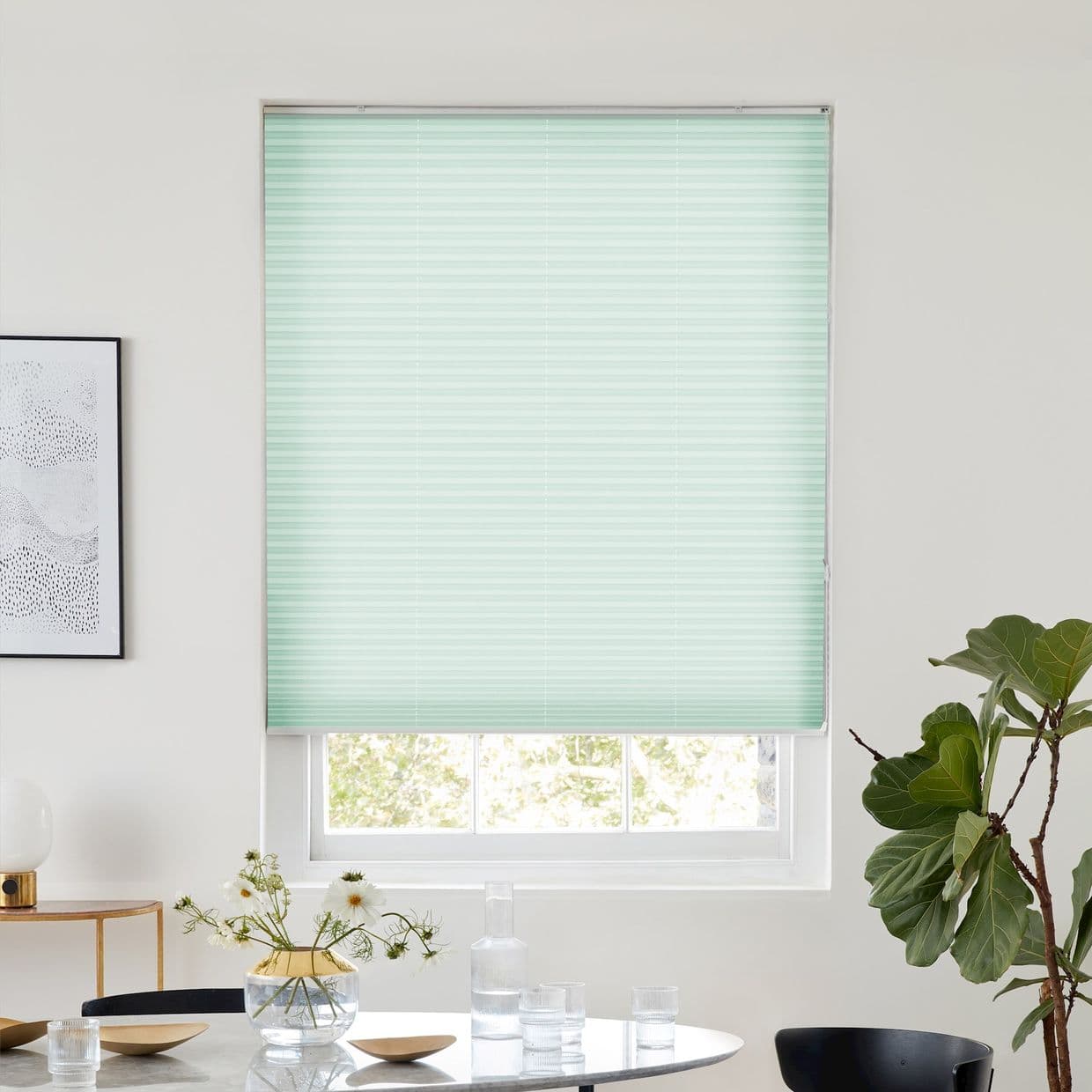 Mint-green pleated blind, fully lowered and filtering daylight; in a white-walled dining nook with round table topped by a glass vase of daisies, carafe and glasses, plus a fiddleâ€‘leaf fig.