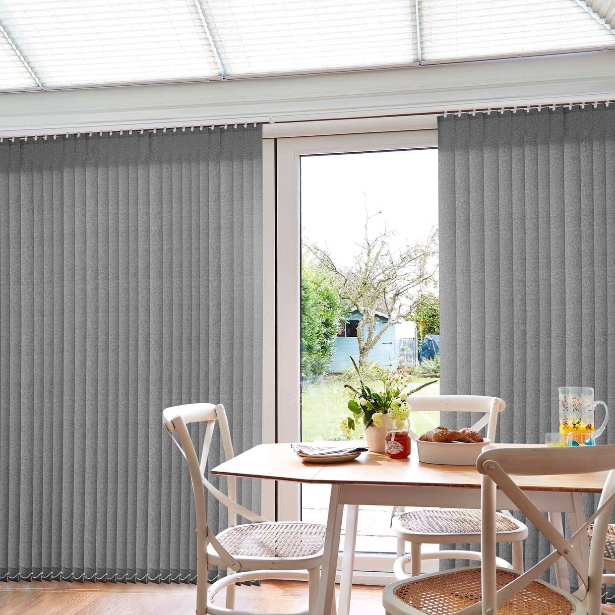 Grey fabric vertical blinds covering sliding glass doors, parted centrally; filtering daylight and revealing a garden view; bright dining area with wooden table, rattan-seat chairs, vase, pastries.