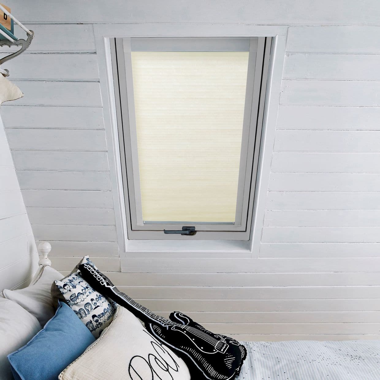 Cream cellular roller blind fully lowered inside a tilted skylight, diffusing soft daylight. Surrounded by white painted tongueâ€‘andâ€‘groove walls above a bed with blue and patterned cushions and guitar cushion.