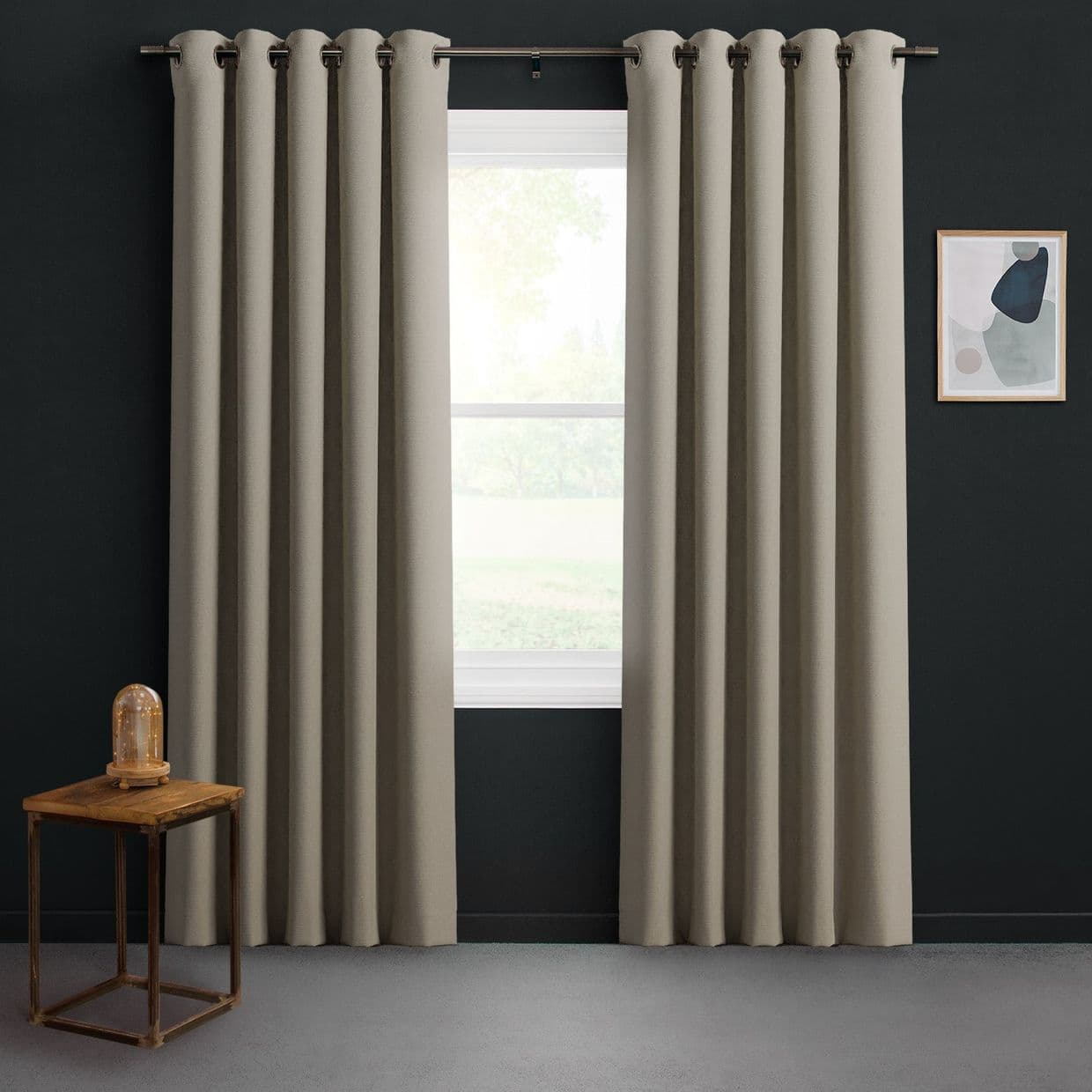 Beige grommet-top curtains, floor-length and pleated, hang on a metal rod, slightly parted to admit diffused daylight; dark charcoal-walled room with a wooden side table, glass-cloche lamp, abstract art.