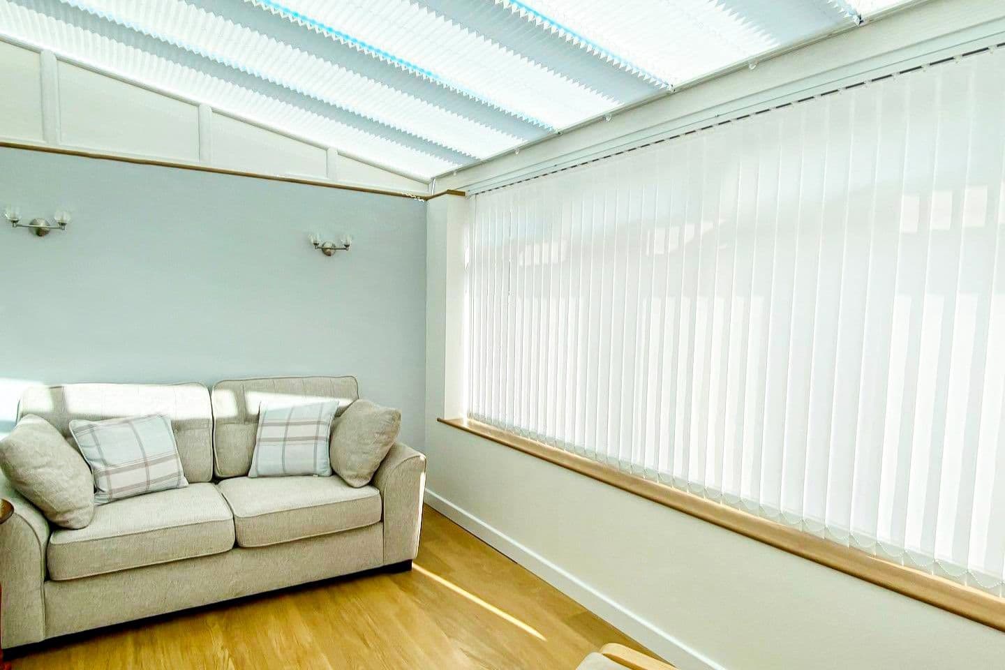 White vertical blinds covering the large side window, fully closed and diffusing bright daylight; pleated roof blinds above. Neutral sunroom with beige sofa, plaid cushions, pale walls and wood floor.