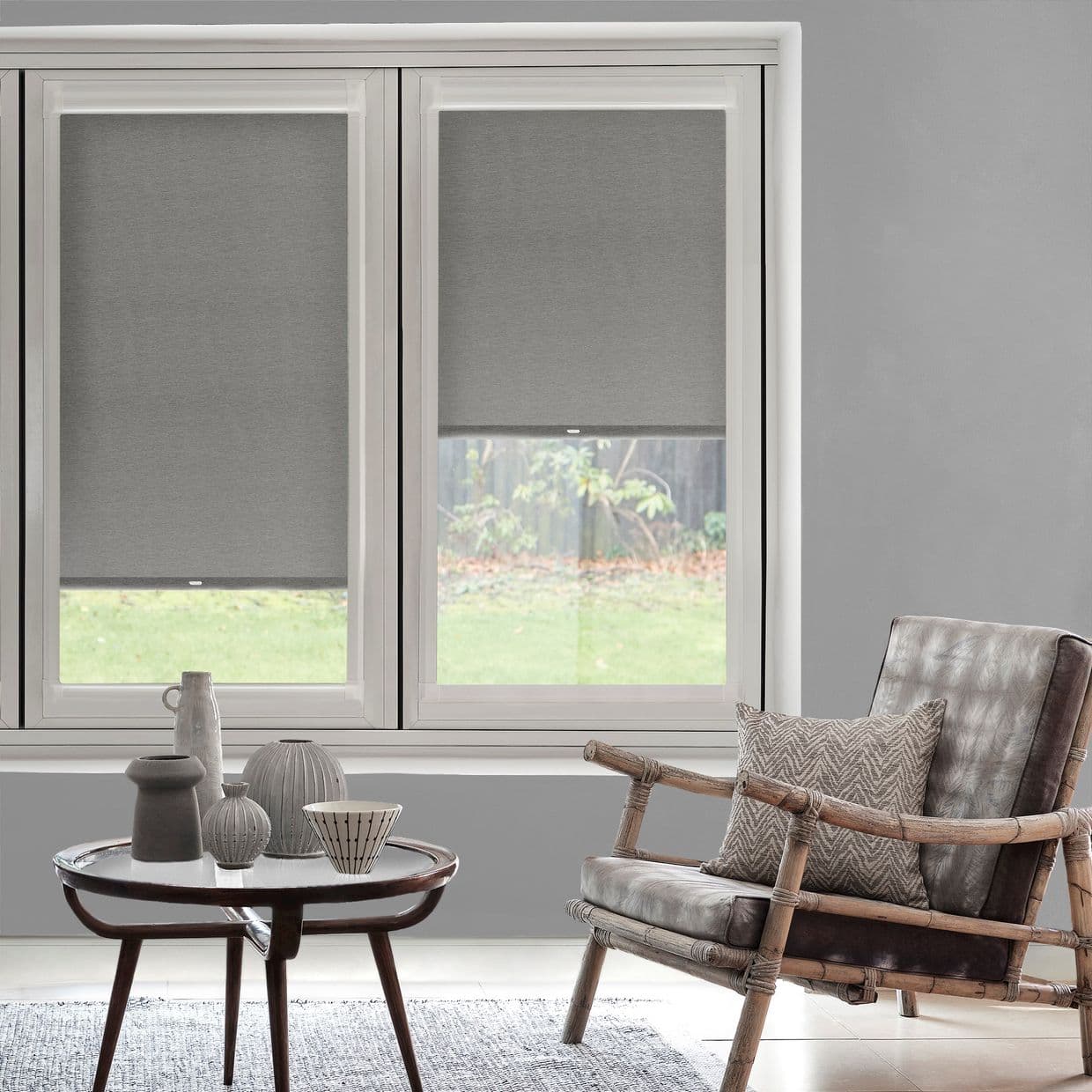 Grey fabric roller blinds covering a two-panel window, lowered to mid-height and filtering light; living room with neutral walls, low coffee table, decorative ceramics, and a rattan armchair.
