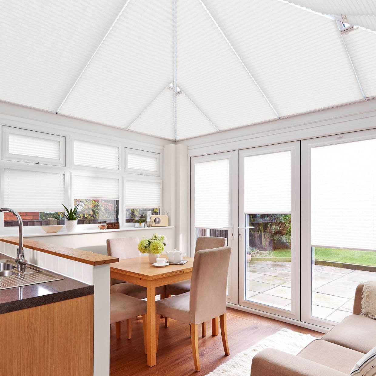 White pleated cellular blinds across the roof, upper windows and French doors; closed, diffusing daylight; in a bright conservatory-style dining/kitchen area with wooden table, chairs and patio view.