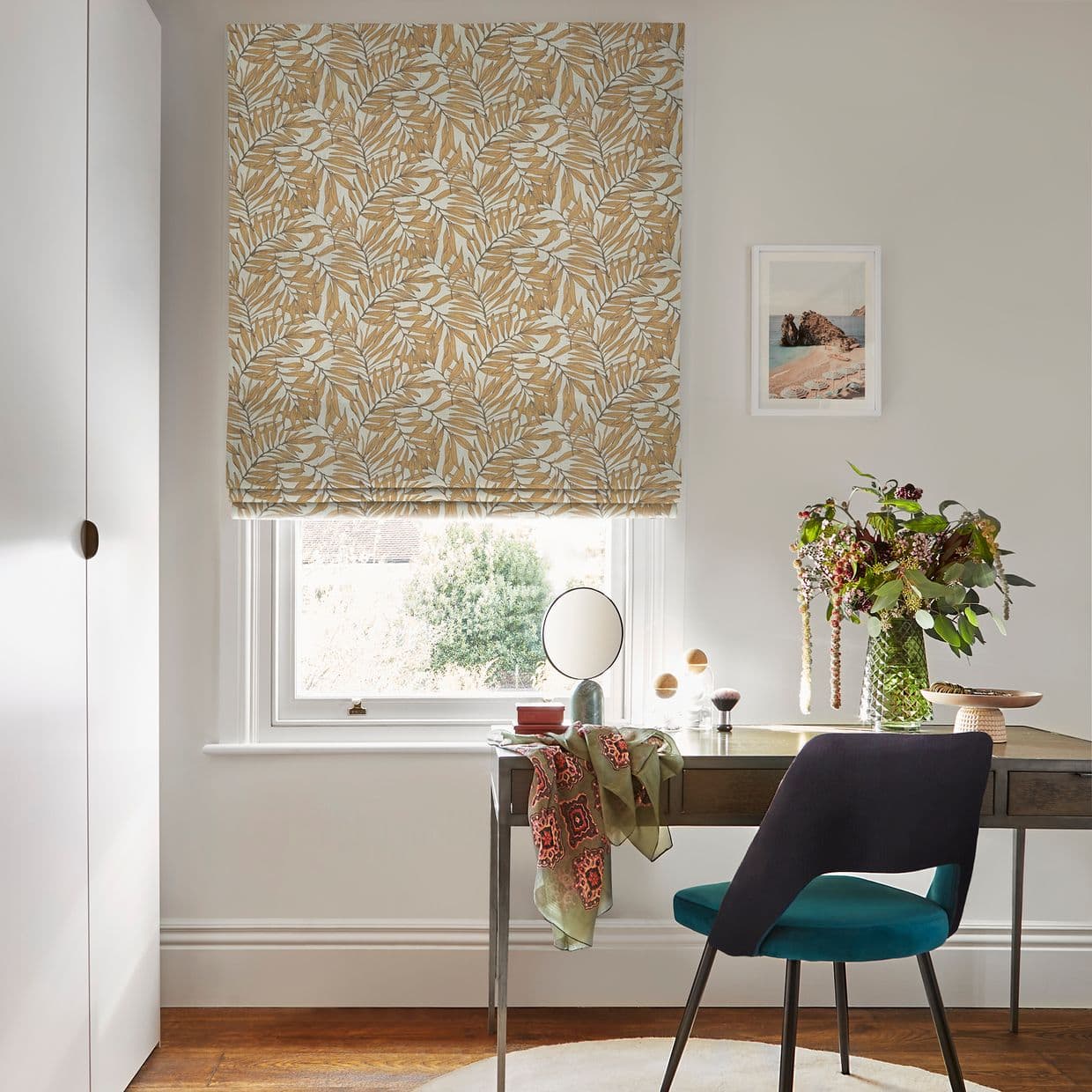 Patterned fabric Roman blind with beige-and-white leaf motif, partially lowered and filtering daylight over a window in a bright home office with desk, teal chair and vase of flowers.