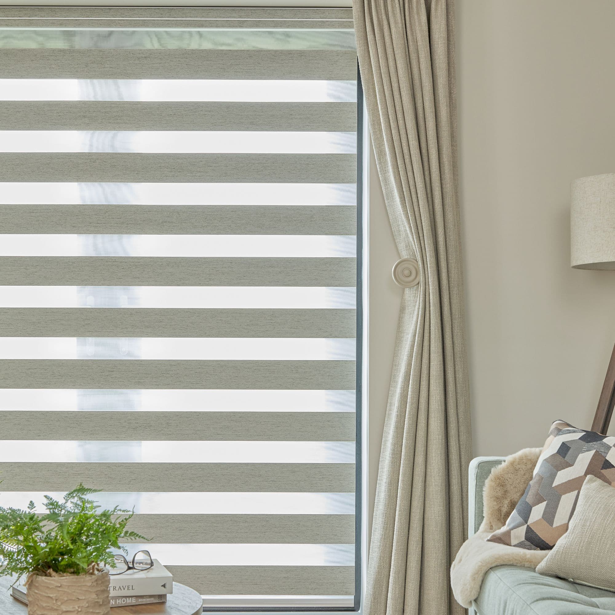 Zebra roller blind with alternating opaque and sheer horizontal bands, lowered and filtering daylight; beige curtain tied back to the right; living-room corner with sofa, lamp, plant, books.

TRAVEL