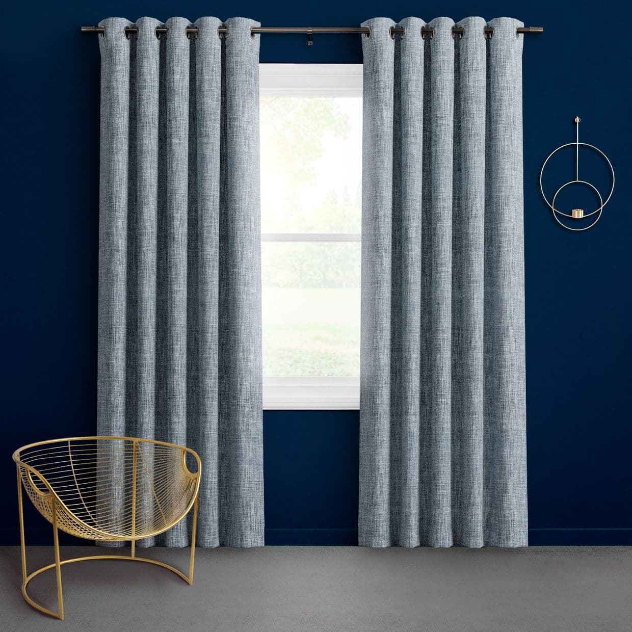 Grey grommet-top curtains, hanging in pleats on a rod and parted to reveal a bright window, against a deep navy wall with a gold chair and round wall decor.