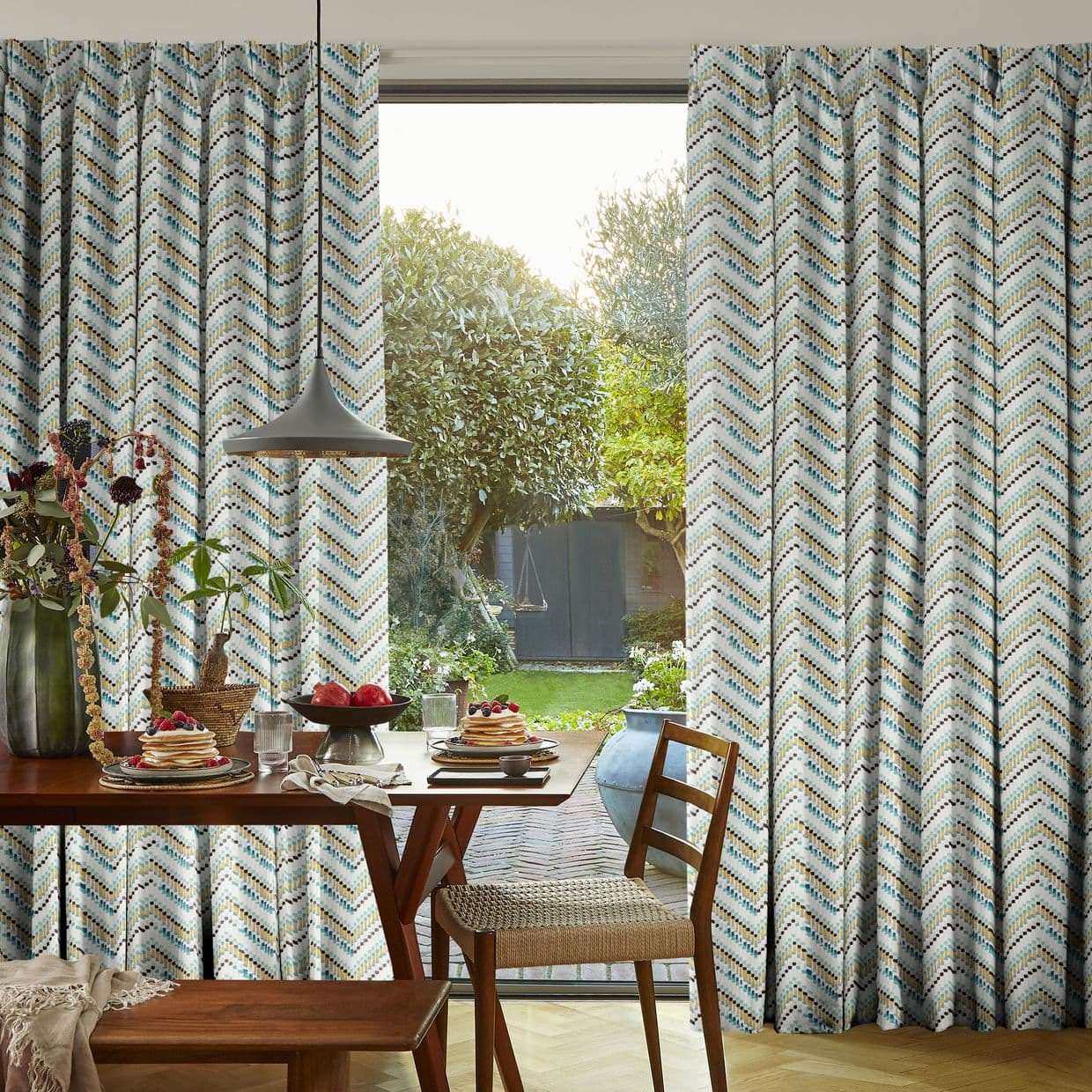 Floor-to-ceiling chevron-patterned curtains hung at a glass door; partially drawn, framing the opening and admitting soft daylight; dining area with wooden table set and a sunlit garden beyond.
