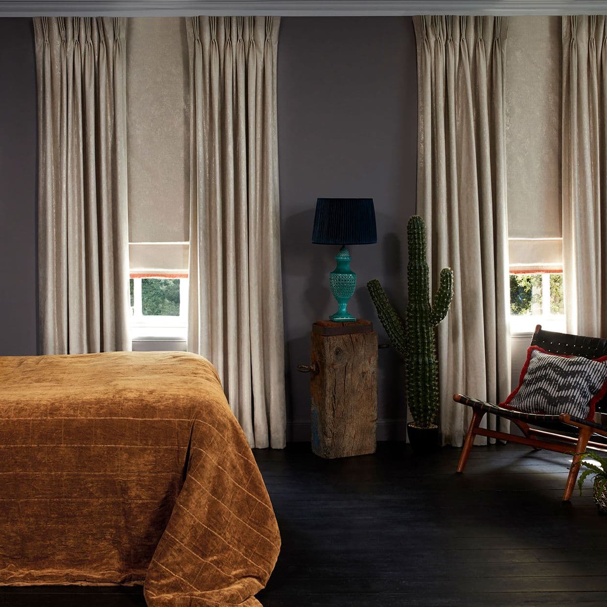 tall beige pleated curtains with lowered Roman shades, partially drawn, softly filtering daylight; bedroom setting with dark floors, rust-colored bedspread, turquoise lamp on a wood stump, potted cactus and chair.
