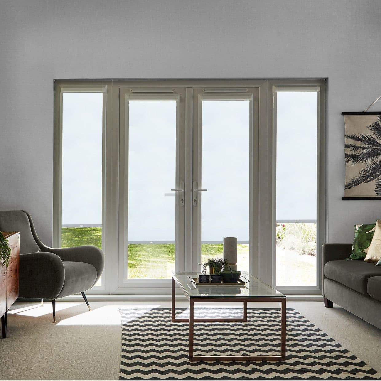 White roller blinds covering tall French doors and sidelights, lowered to mid-height and filtering daylight; modern living room with gray seating, glass coffee table, chevron rug, and lawn visible outside.