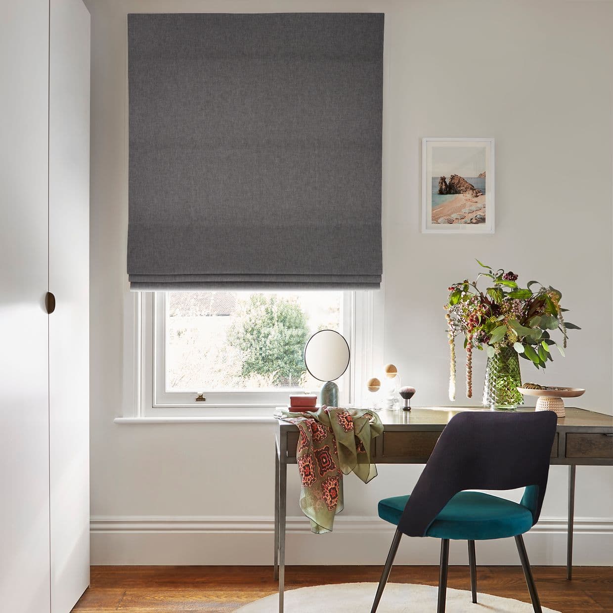 Grey fabric Roman blind, lowered above the sill with neat horizontal folds, filtering daylight over a desk with teal chair, vase of flowers, mirror and framed seaside print, garden beyond.