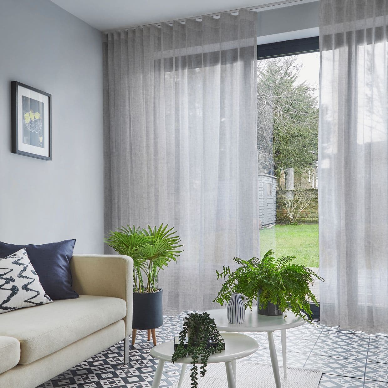 Sheer grey floor-to-ceiling pleated curtains, partly drawn and filtering daylight, revealing a gap to a lawn and shed; modern living room with pale sofa, patterned tile floor and potted plants.