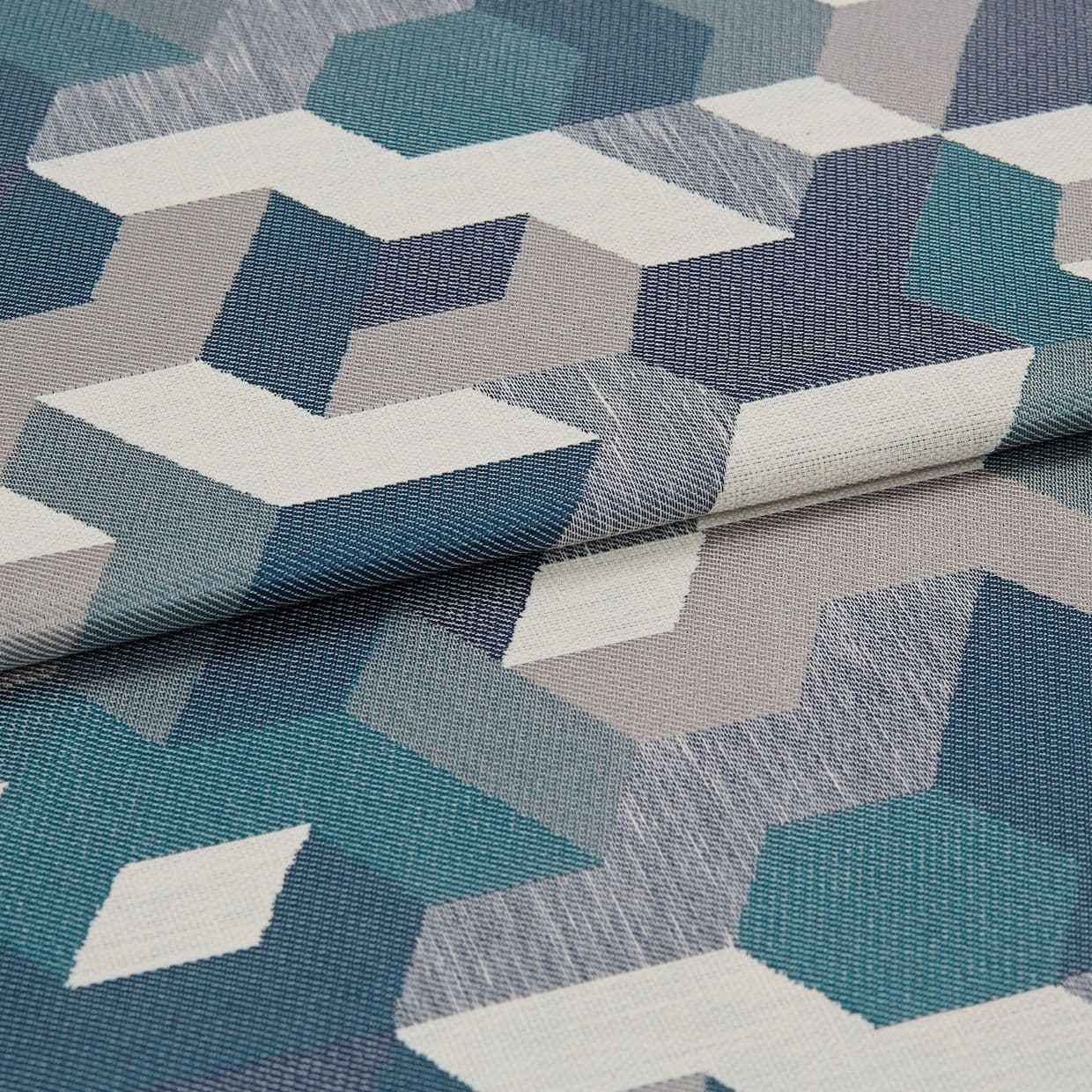 Woven upholstery fabric with teal, navy, gray and white isometric geometric pattern â€” folded once, overlapping â€” close-up on a flat surface under soft, neutral lighting.