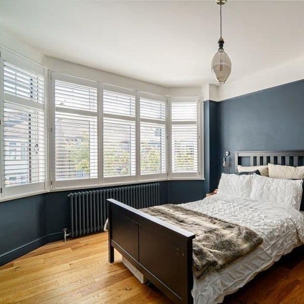 White plantation shutters on a bay windowâ€”five slatted panels with adjustable louvers partially openâ€”filter soft daylight into a bedroom with dark blue walls, hardwood floor, black bed, and radiator.
