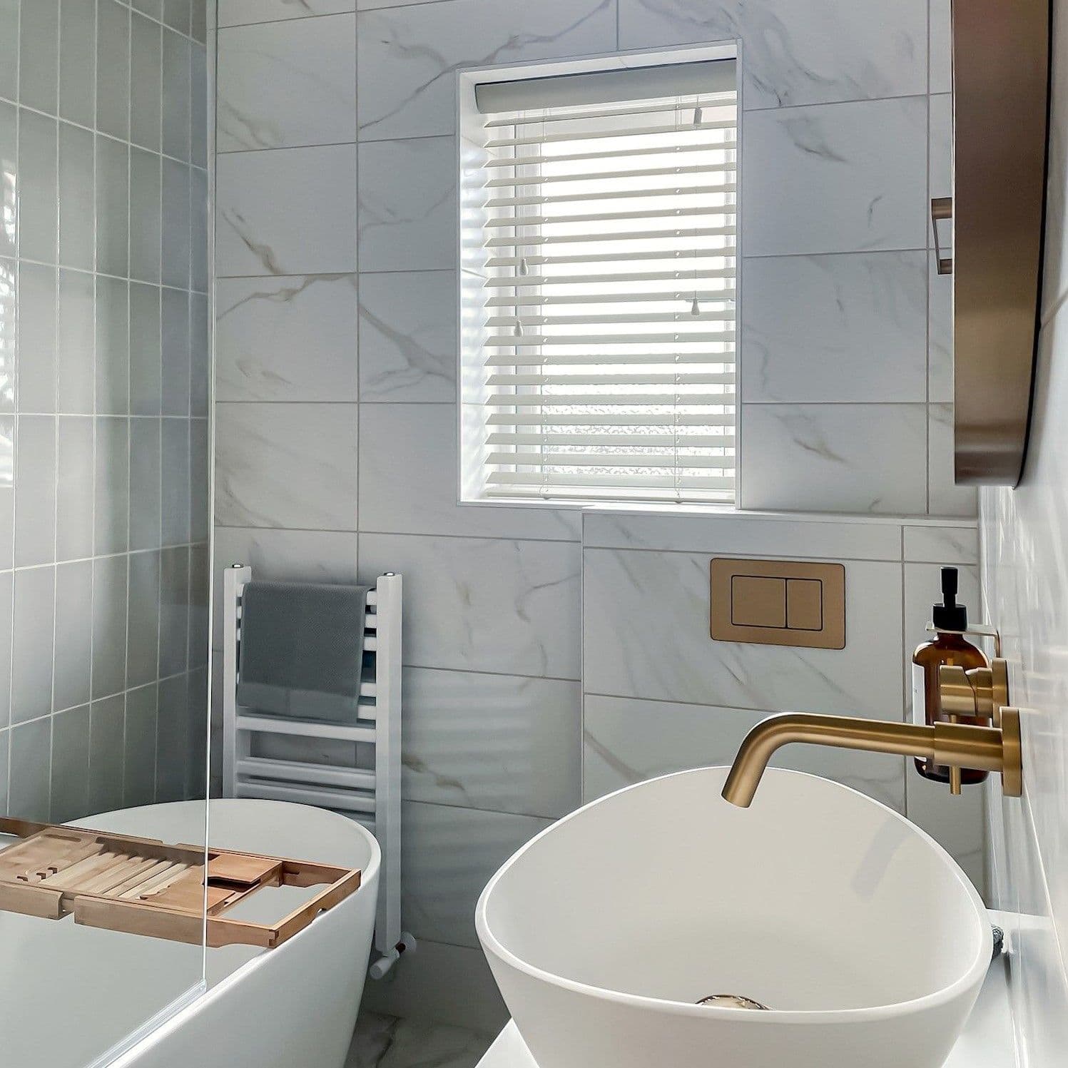 White horizontal blinds in a recessed window, partially closed and filtering daylight, in a marble-tiled bathroom with freestanding tub, bath tray, vessel sink, brass faucet and towel rail.