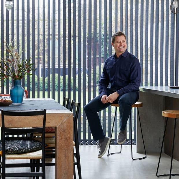 Floor-to-ceiling vertical blinds with narrow dark slats, mostly closed yet permitting garden views and daylight; modern kitchen/dining interior with wooden table, vase, man sitting on barstool smiling.