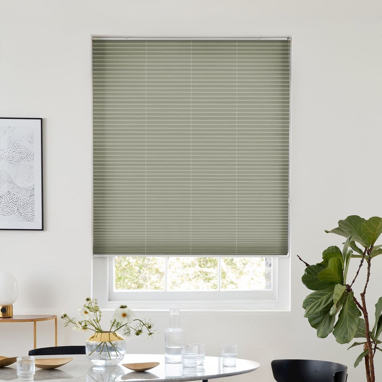 Sage-green pleated blind, fully lowered over a window with visible lower sash, filtering daylight; above a dining table with glassware, vase of daisies, framed art and a fiddleâ€‘leaf fig.
