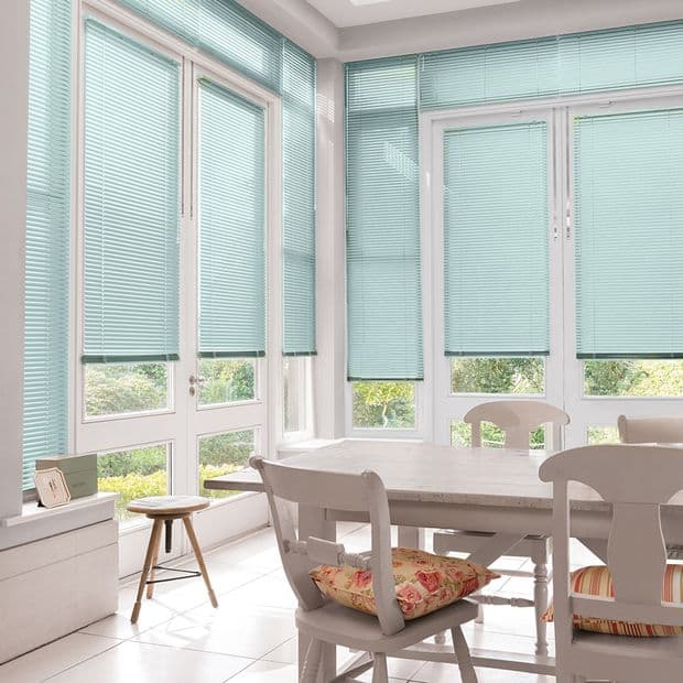 Turquoise horizontal Venetian blinds covering tall bay windows and glass doors, lowered and slightly tilted to filter soft daylight; dining area with white wooden table and chairs, garden visible outside.