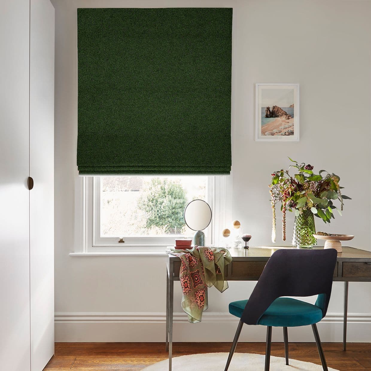 Dark green Roman blind, fully lowered with neat horizontal folds, blocking light; bright home office with wooden desk, teal chair, vase of flowers and outside garden visible.
