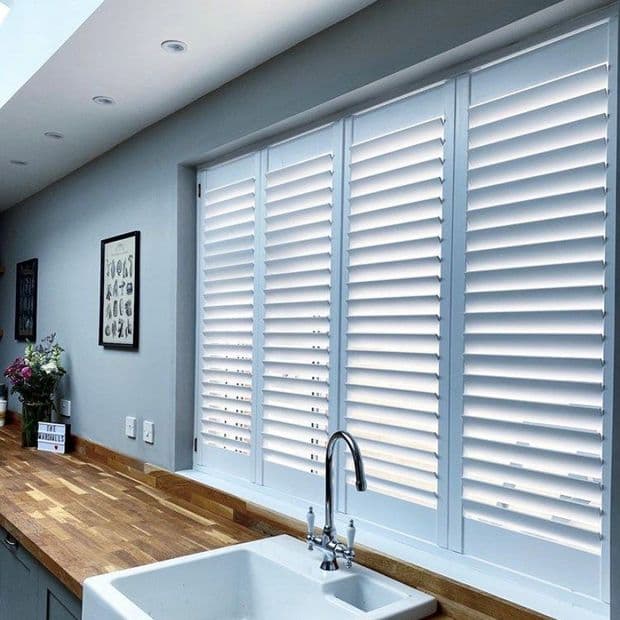 White plantation shutters covering a wide kitchen window, slats tilted to diffuse daylight; farmhouse sink and chrome faucet beneath; wooden countertop, muted grey walls and soft natural light.