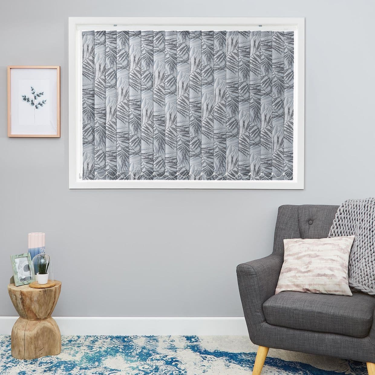 Fabric vertical blinds with a grey fern-leaf pattern, fully closed across the window, diffusing daylight; set in a pale-grey living room beside a grey armchair, wooden side table, and blue rug.
