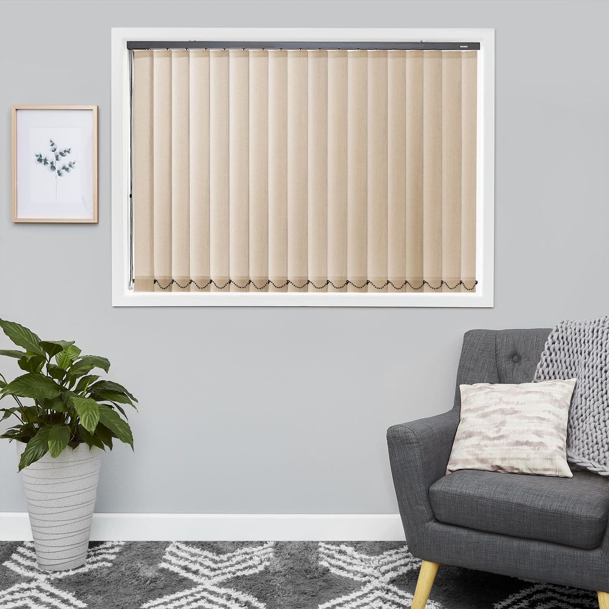 Beige vertical blinds, evenly spaced slats with bottom bead chain, fully closed over a white-framed window, softly filtering daylight in a gray living room with armchair, plant, framed botanical print.