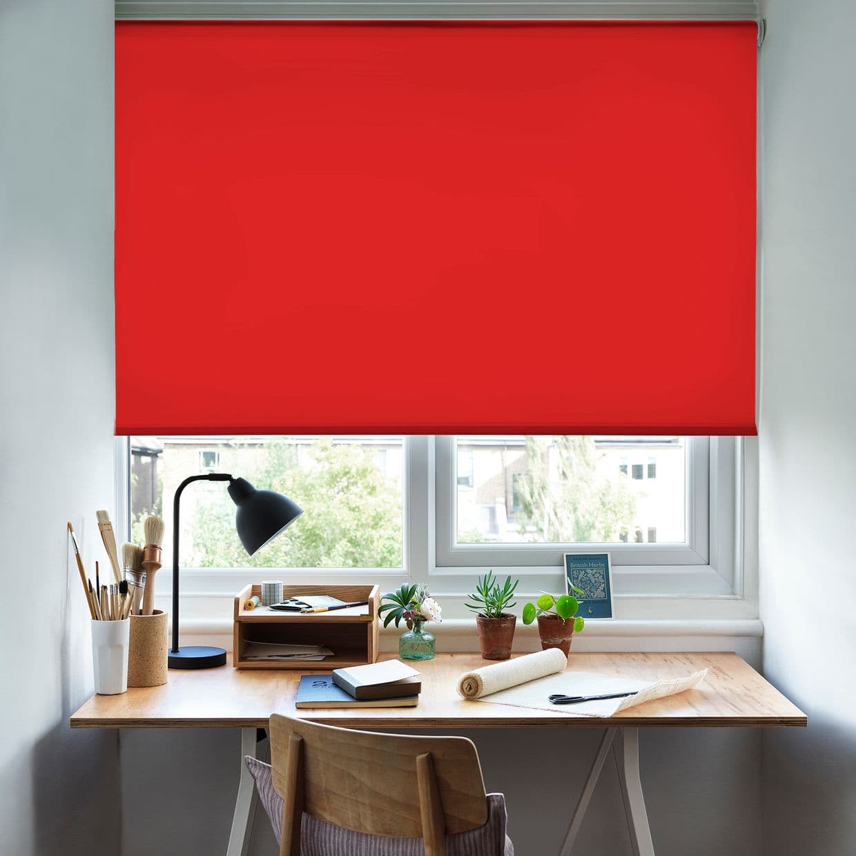 Red roller blind, fully lowered and opaque, blocking daylight over a tidy home desk with lamp, potted plants, art brushes, and a window's soft natural light around the edges.