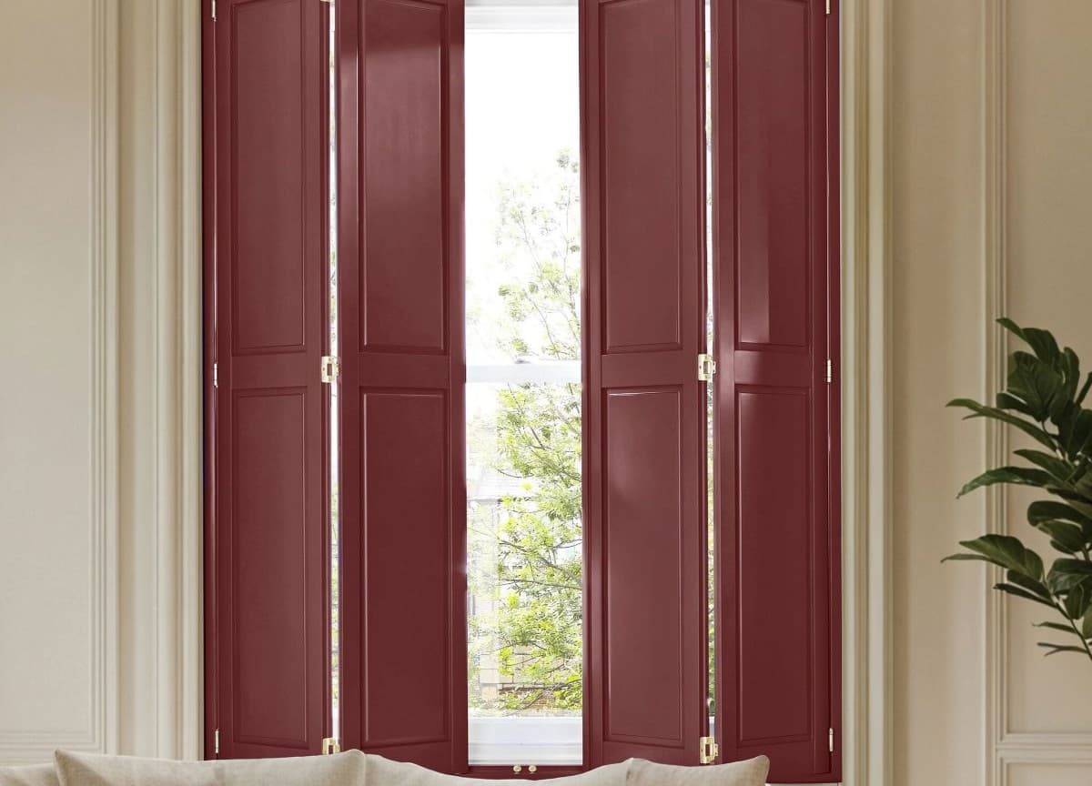 Burgundy four-panel interior folding shutters, raised-panel hinged, largely closed with a narrow central gap revealing daylight and tree branches; beige moulded living room with sofa cushion and potted plant.