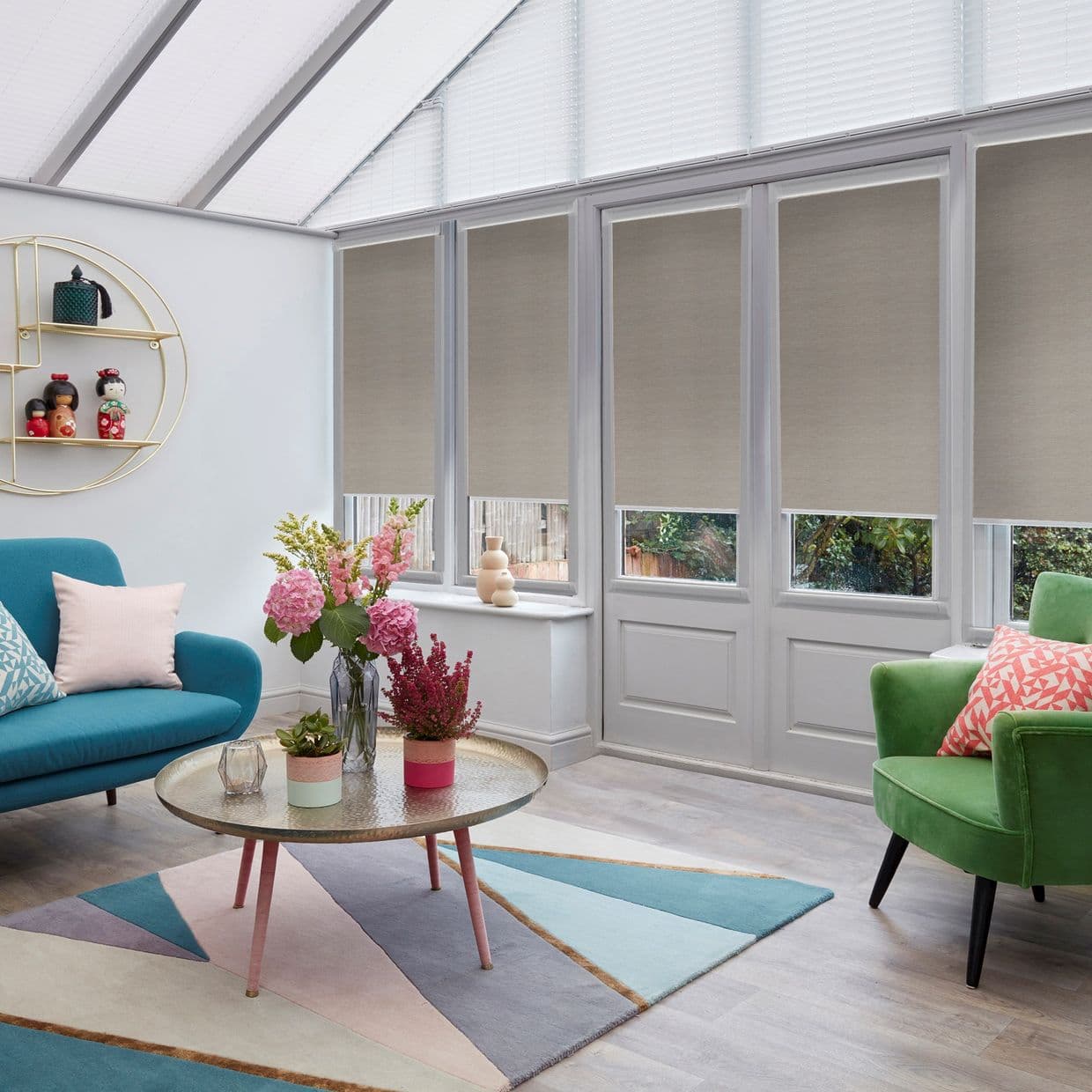 Beige roller blinds lowered to mid-height across tall conservatory windows, filtering light; room features glass-pitched roof, teal sofa, green armchair, pastel geometric rug and a round coffee table with flowers.