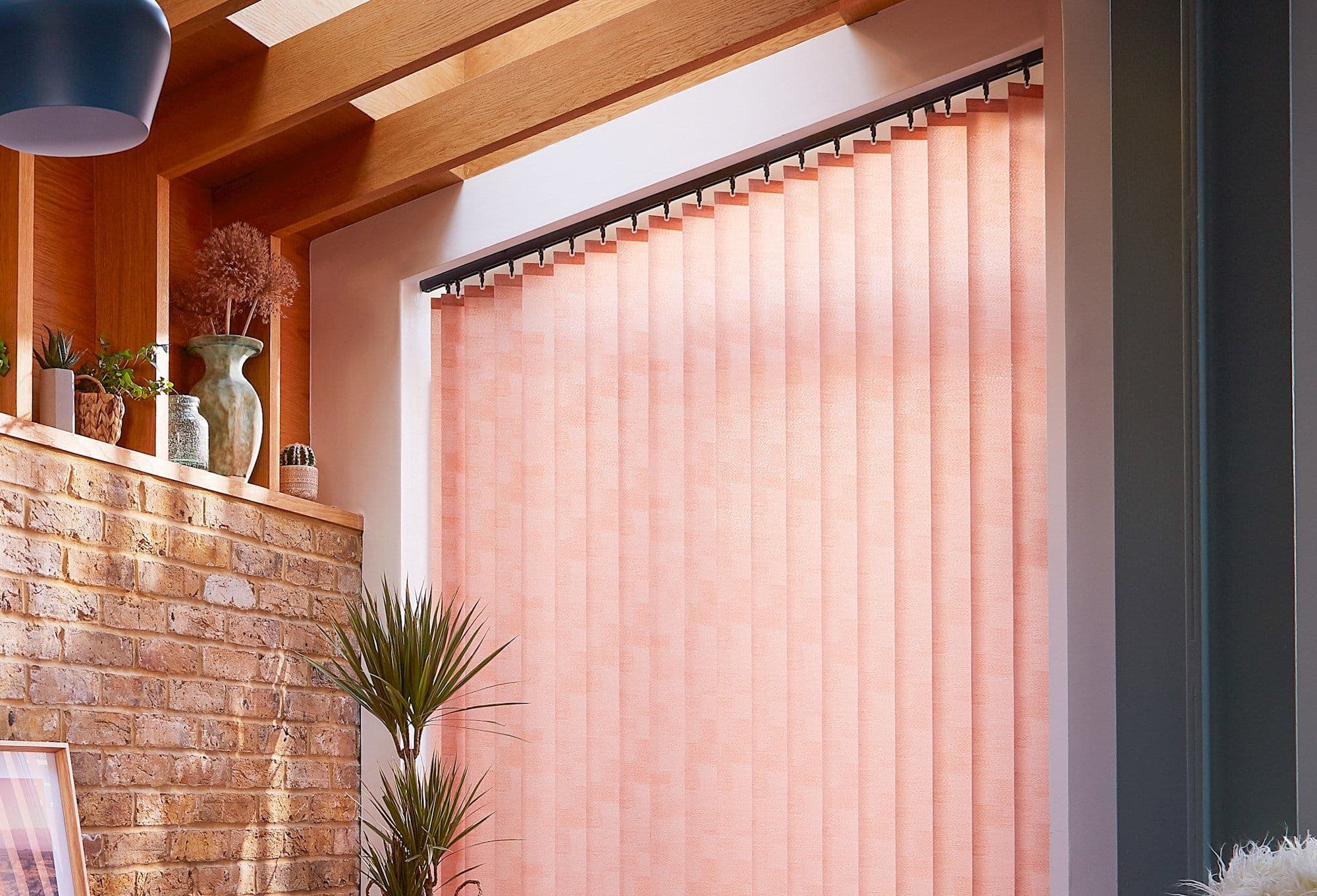 Peach textured vertical blinds, hung on a slanted track, closed and softly diffusing light, in a warm living space with exposed brick, wooden beams, potted plants, and a skylight.