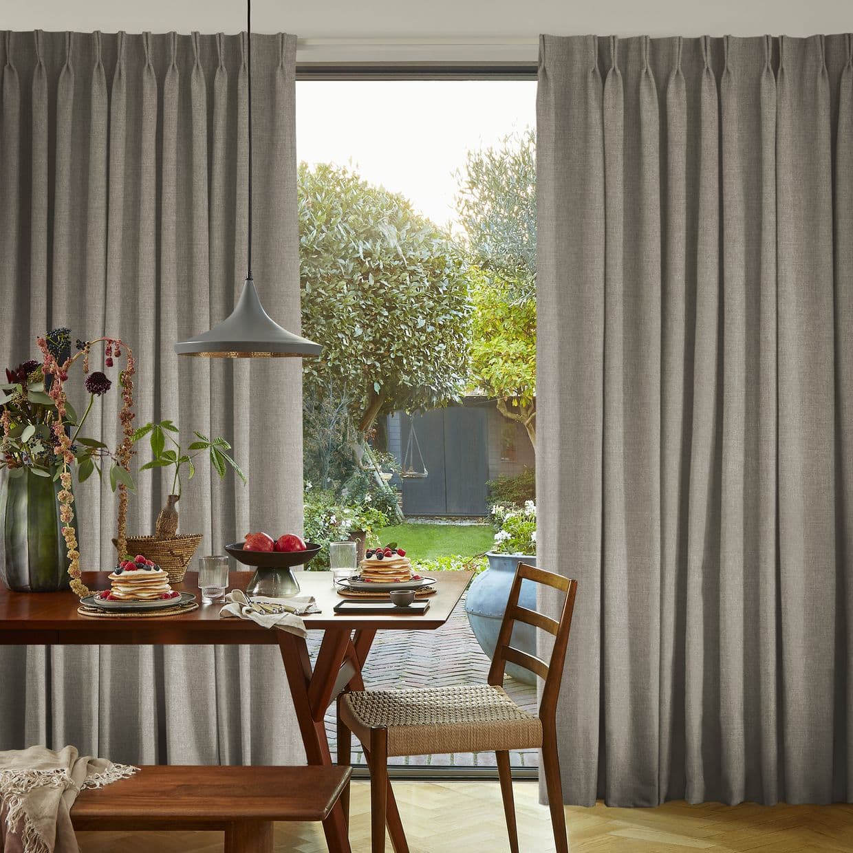 Full-length pleated gray curtains, drawn aside revealing a central glass door; filtering morning light into a dining area with wooden table set for breakfast, a pendant lamp, and a garden view.
