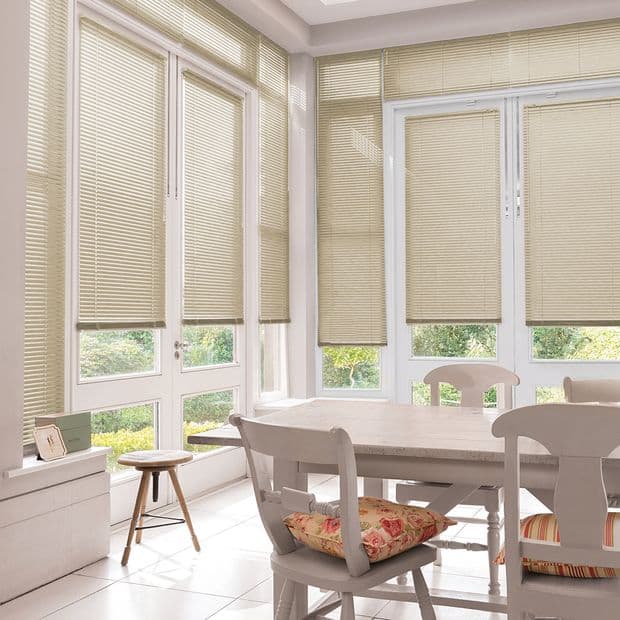 Beige horizontal Venetian blinds covering tall French-style windows and doors, partially lowered, filtering soft daylight into a bright dining area with white table, wooden chairs and floral cushions, garden view.