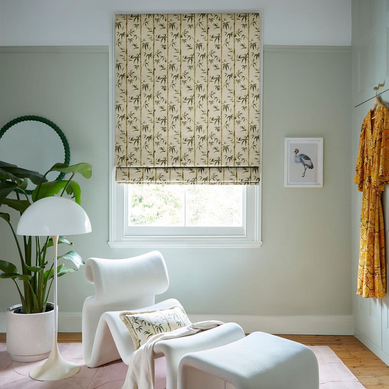 A beige Roman blind patterned with vertical bamboo, lowered over the window and softly filtering daylight, in a pale green room with white lounge chair, plant, lamp and bird print.