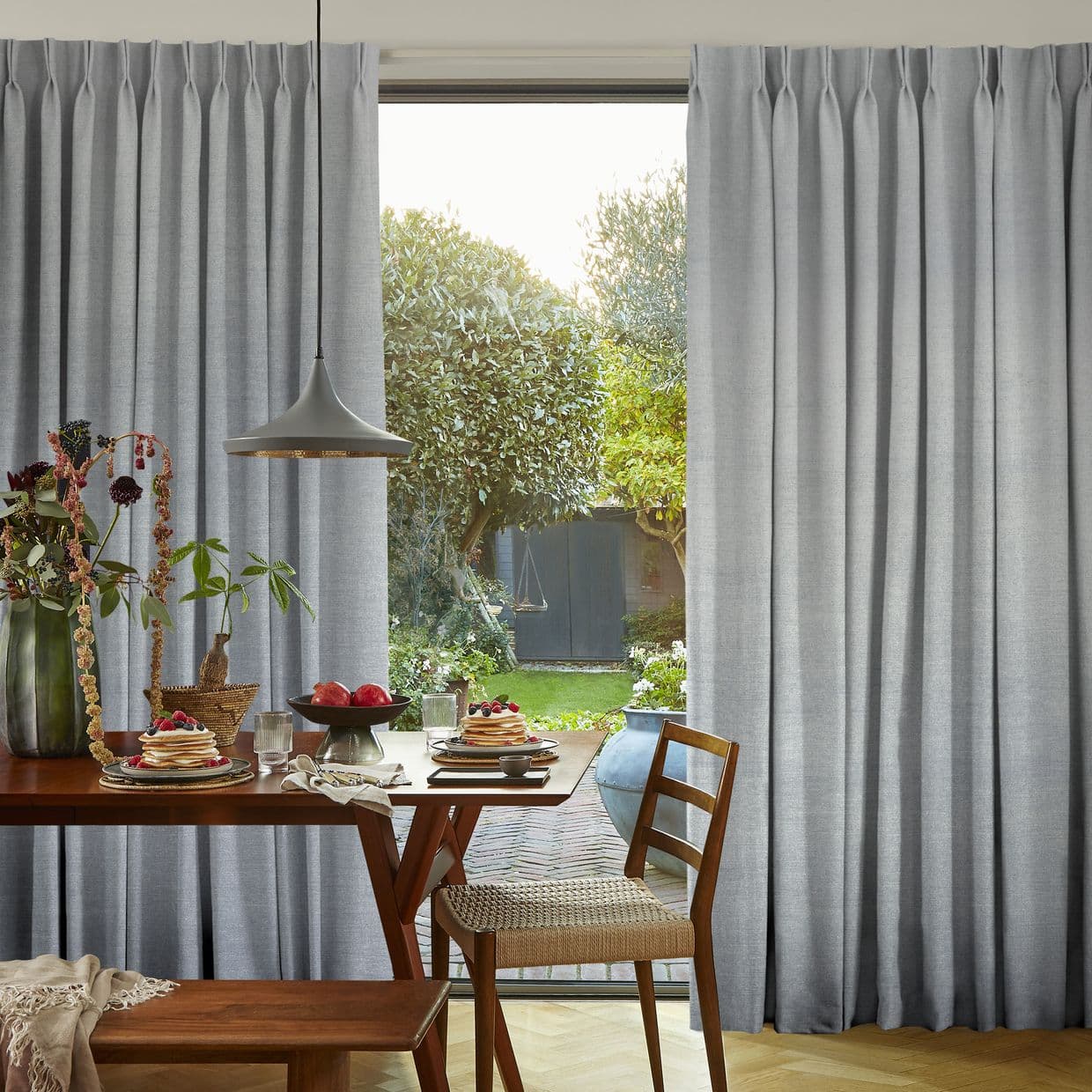 Pleated floor-length grey curtains, drawn open at the center, framing and filtering daylight through a glass door into a dining area with wooden table, pendant lamp, and sunlit garden beyond.