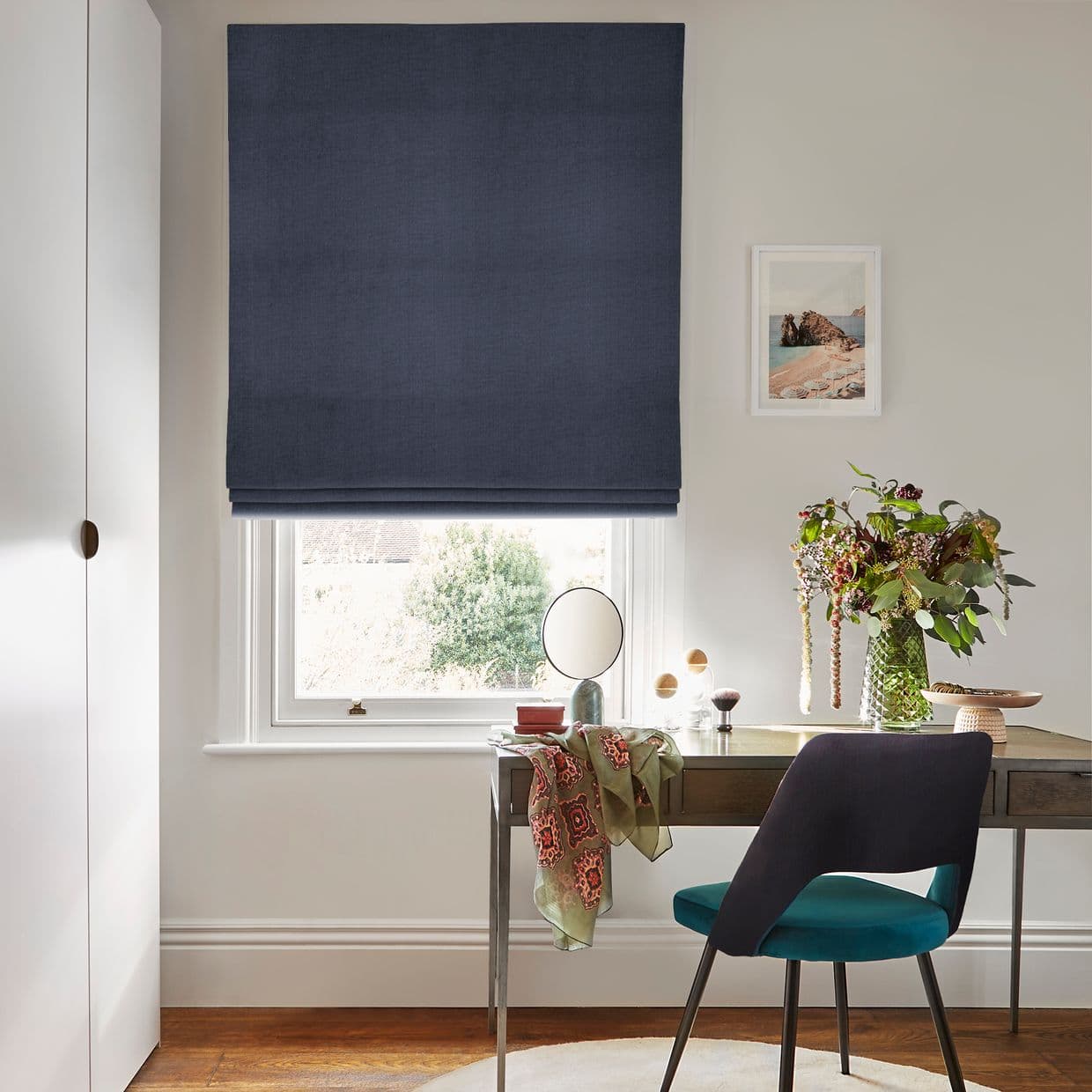 A large navy Roman blind, fully lowered with soft horizontal folds, filtering daylight through a small lower gap, in a bright home workspace with desk, teal chair, vase of flowers.