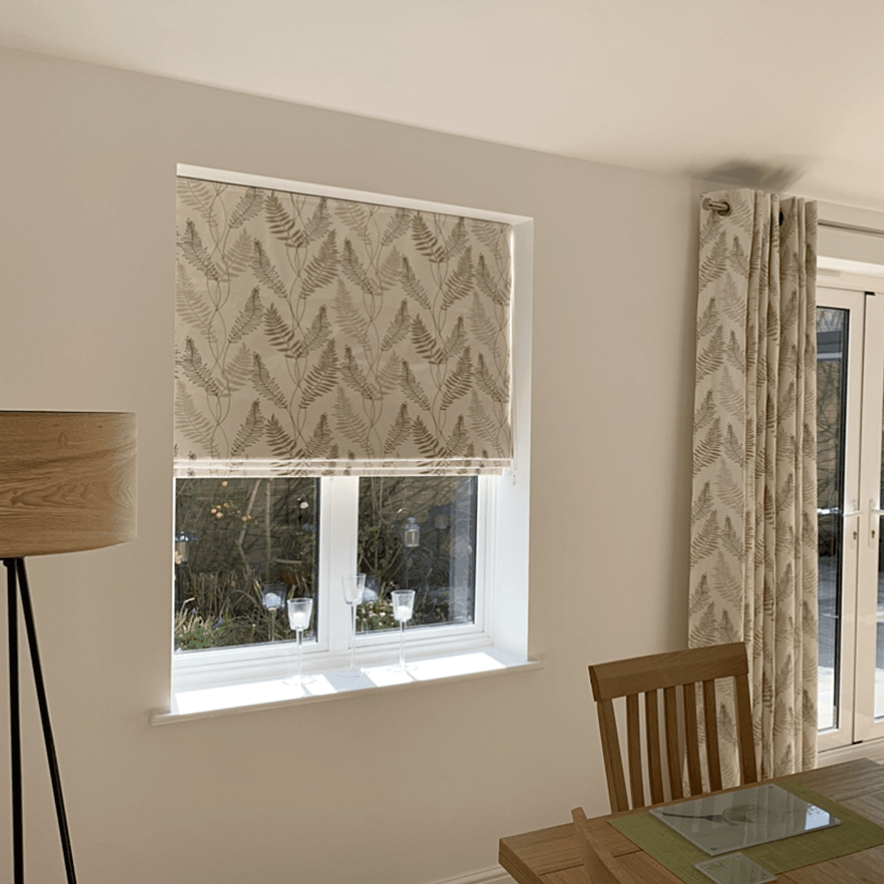 Patterned roller blind and matching grommet-top curtain, beige with fern print, lowered over a sash window (blind half-down) filtering daylight; dining area with wooden table, chair, lamp and garden view.