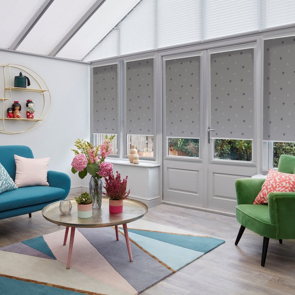 Grey star-patterned roller blinds over conservatory French doors and windows, partially lowered and filtering daylight, in a bright sunroom with glass roof, colorful sofas, rug and round coffee table.