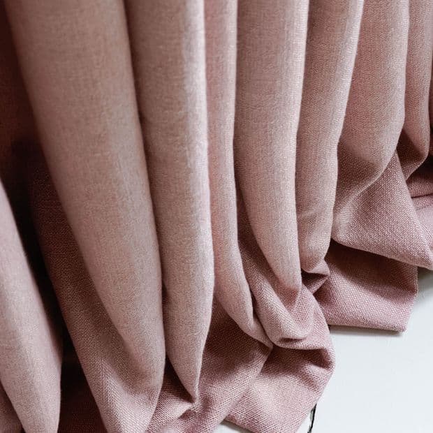 Textured pink curtains hang in deep vertical pleats, pooling on the floor; soft folds filter diffuse light, suggesting a softly lit interior space.