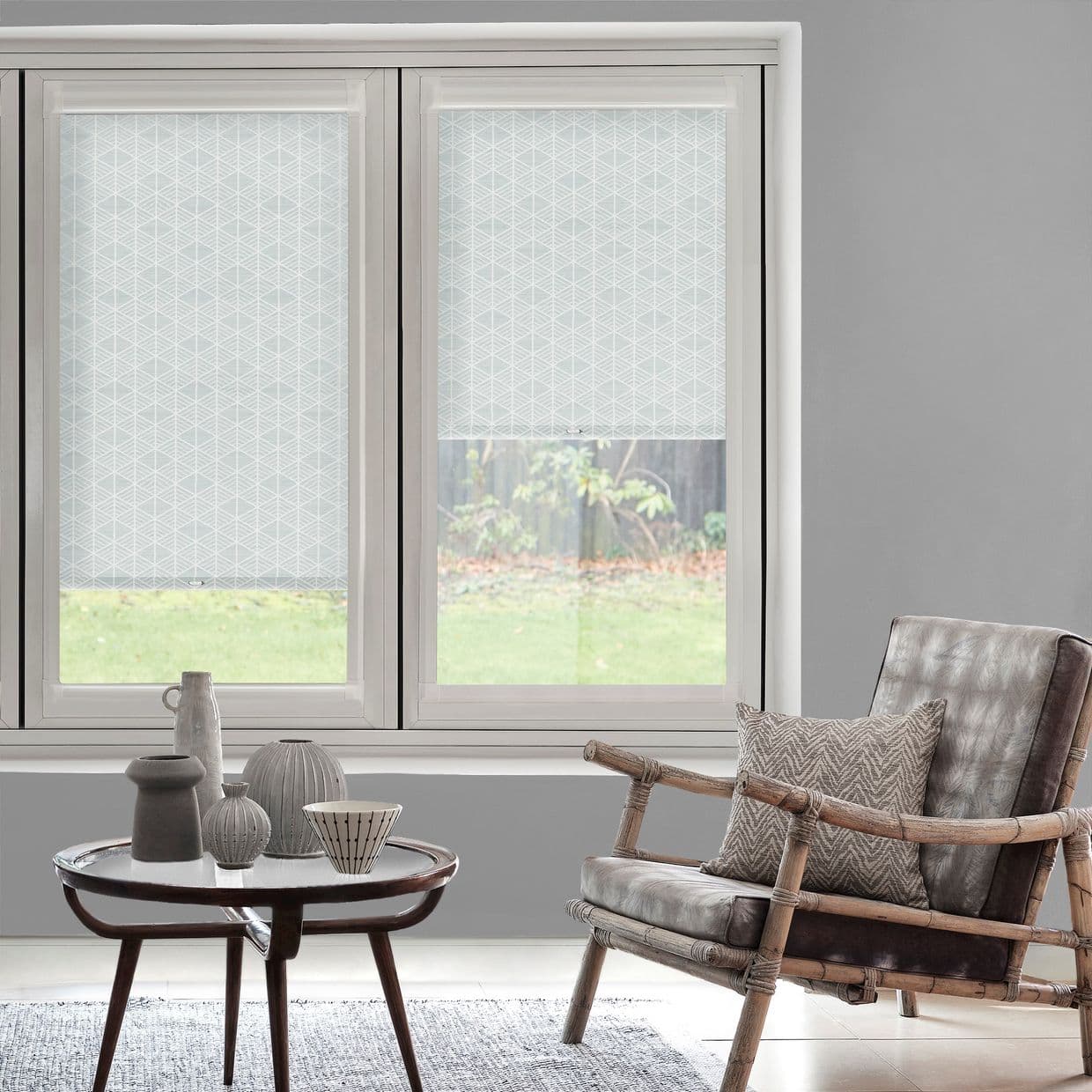 Patterned roller blinds on two adjacent windows, partially lowered with pale geometric print and transparent bottoms; softly lit living room with rattan armchair, coffee table, and lawn view.