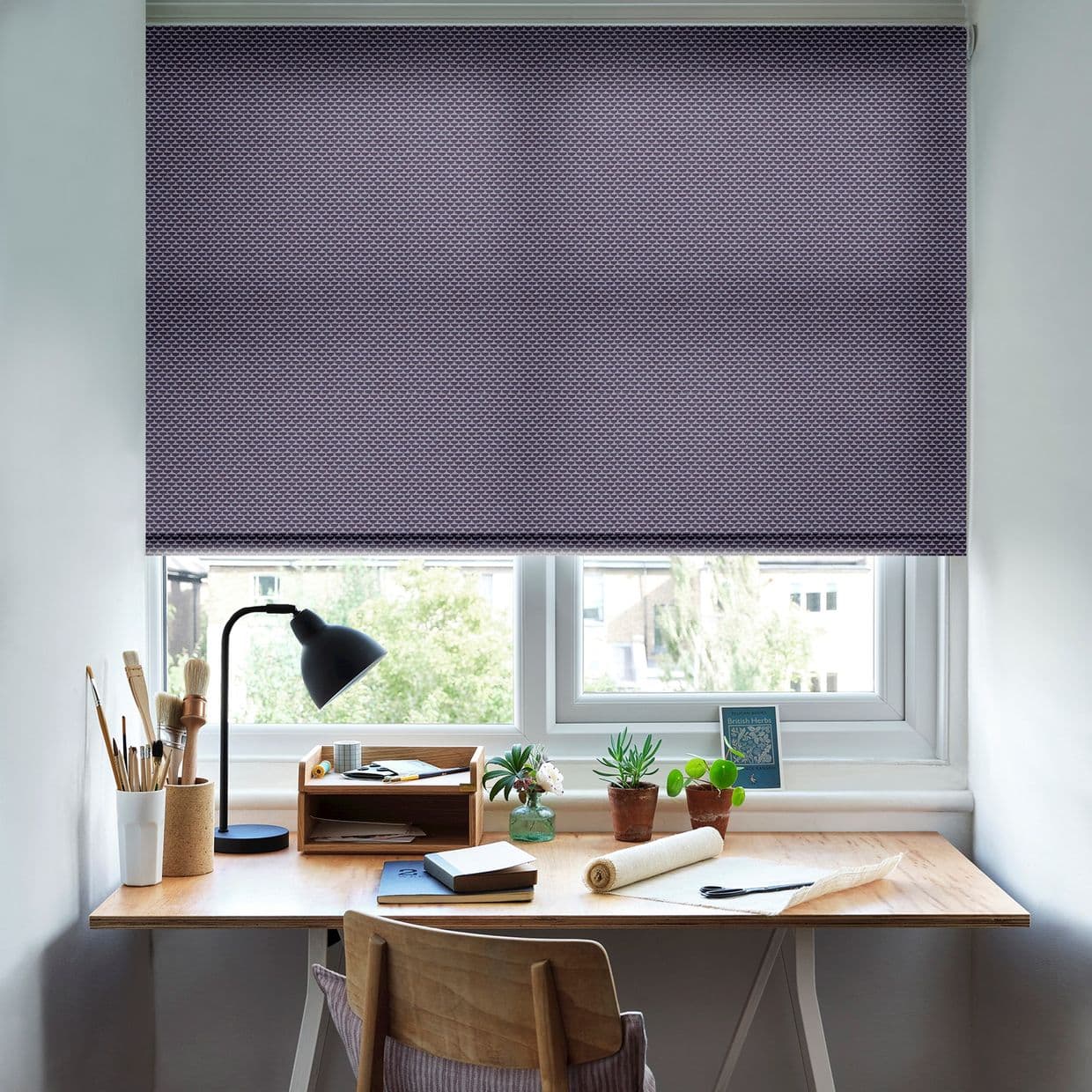 Purple patterned roller blind, lowered nearly to the sill, filtering daylight; a wooden desk beneath holds a black task lamp, brushes, notebooks and small potted succulents by the window.