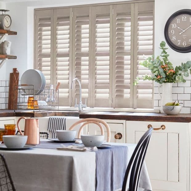 Beige louvered plantation shutters covering a wide kitchen window, slats partially tilted filtering soft daylight; framed by a sink and wooden countertop, dining table set, subway-tile backsplash and plant.