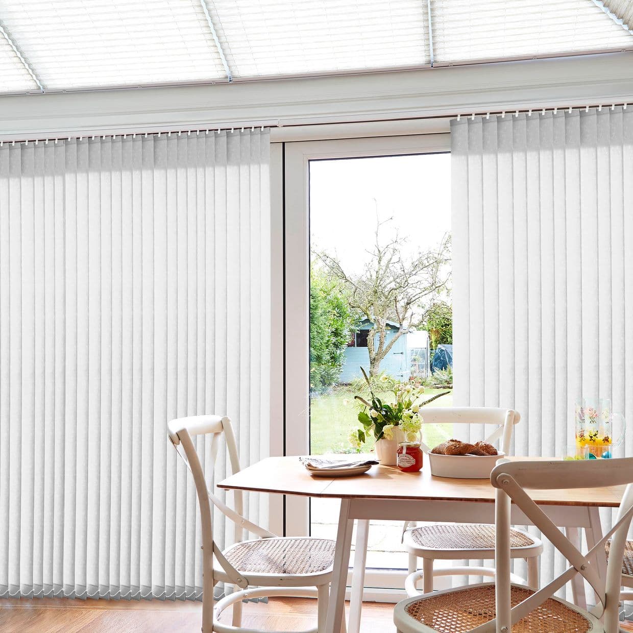 White vertical blinds covering glass doors, mostly closed with a central gap; filtering daylight and parted to reveal a backyard tree and shed beyond a dining table with chairs.