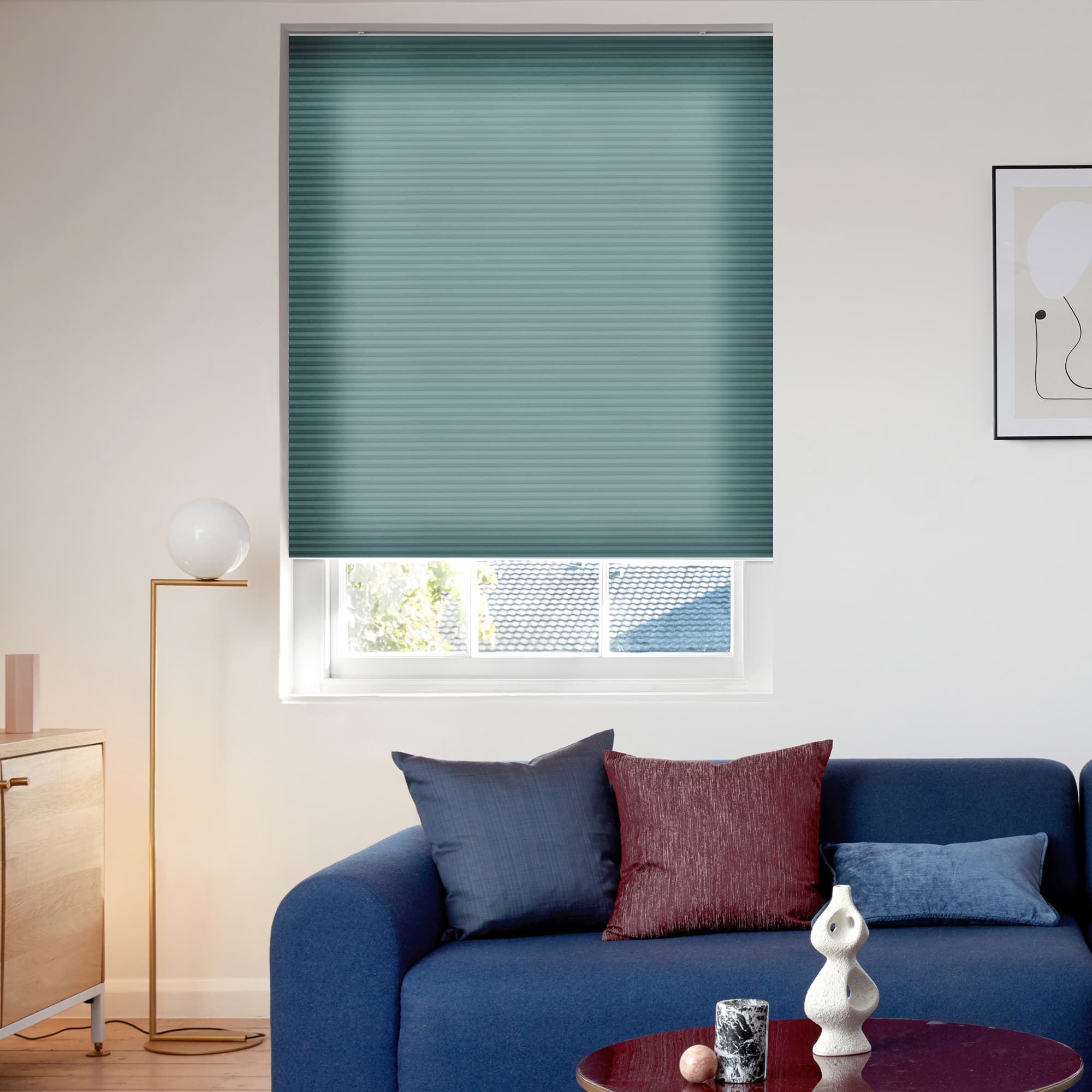 Teal pleated cellular blind, fully lowered with a narrow bottom reveal; filtering daylight and partially blocking a roof view â€” above a blue sofa in a modern, sunlit living room.