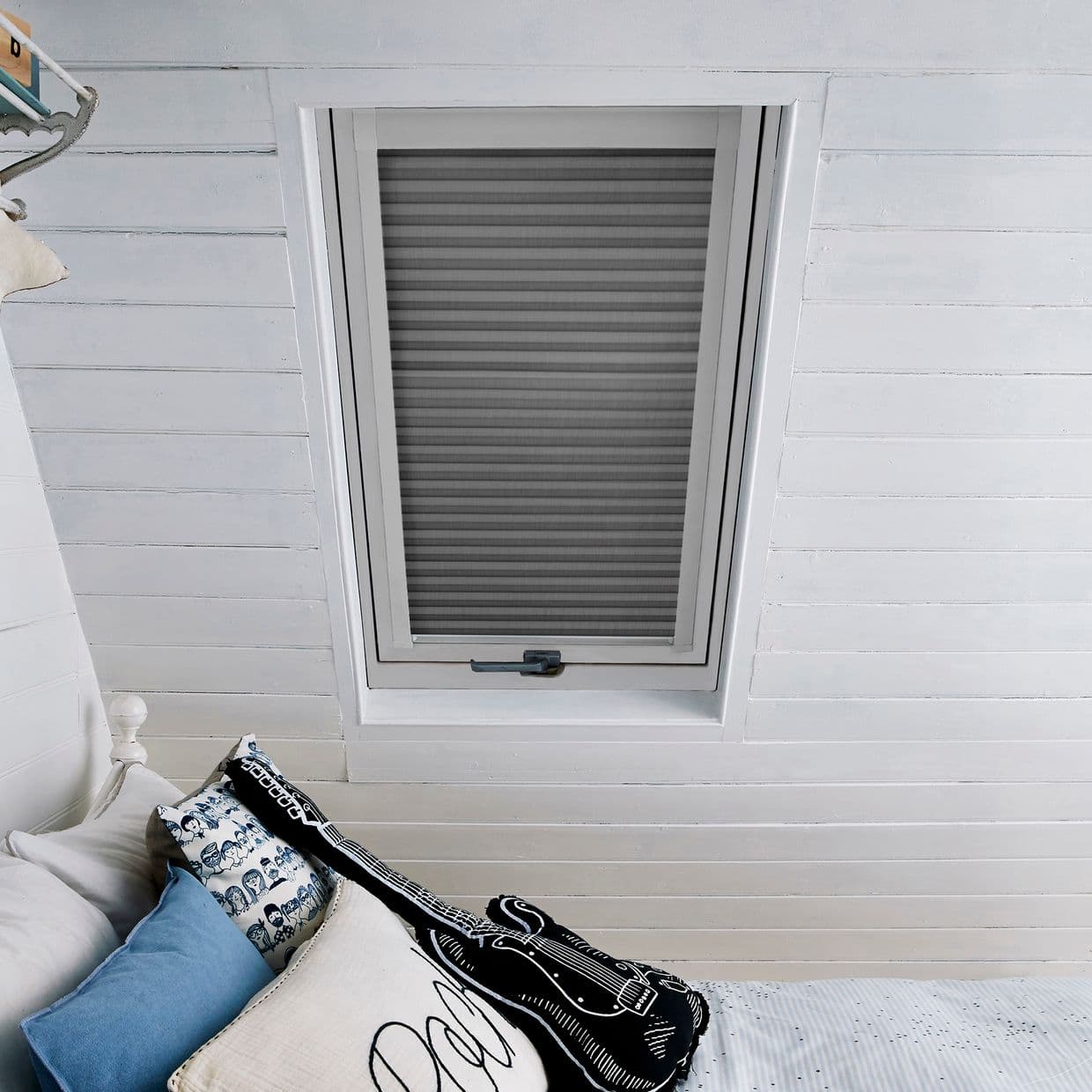 White pleated cellular blind in a roof skylight, fully lowered with horizontal folds, filtering daylight; set into white-painted tongue-and-groove ceiling above a bed with patterned cushions and a guitar cushion.