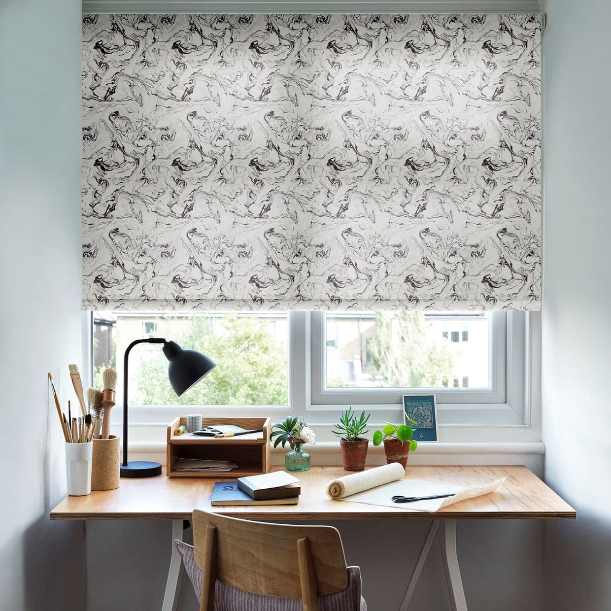 Marbled roller blind, lowered and filtering daylight; covering a double-pane window above a tidy wooden desk with lamp, art brushes, notebooks and potted plants in a bright home workspace.