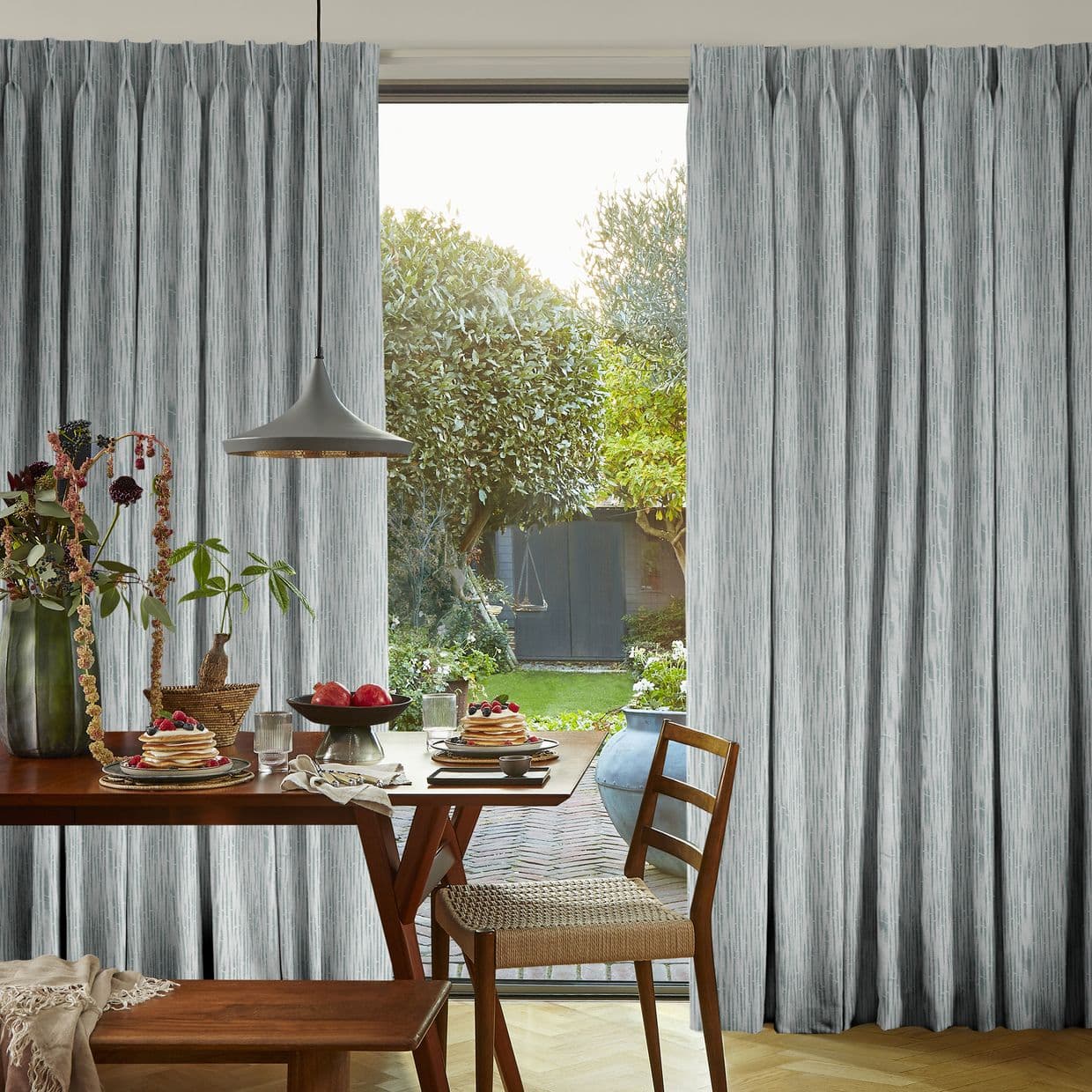 Floor-to-ceiling pleated grey curtains, hanging on either side of a sliding glass door, parted to reveal a sunlit backyard; interior dining table set with pancakes, pendant light and woven chair.